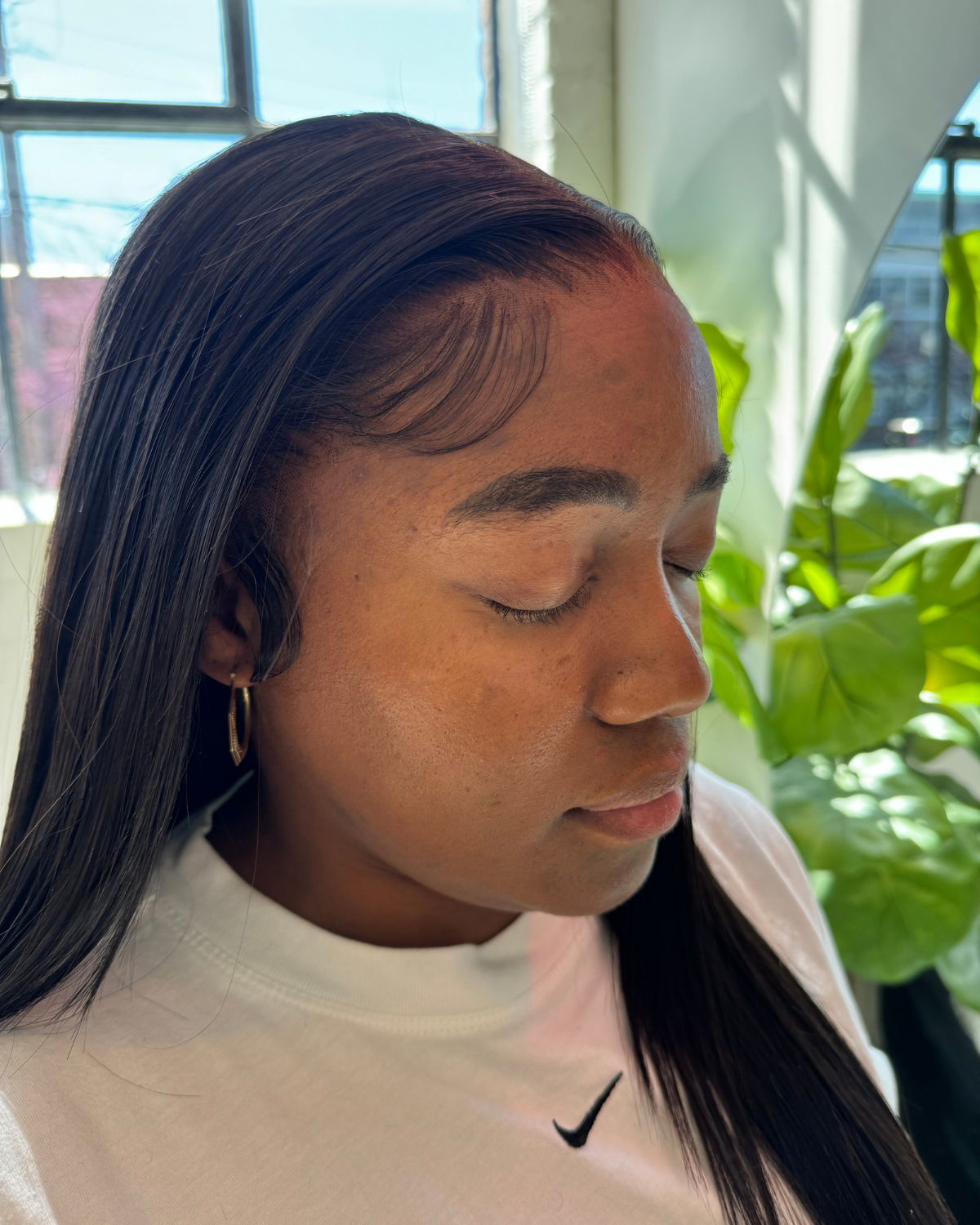 Person wearing a white Nike shirt and a frontal medical wig with plants in the background
